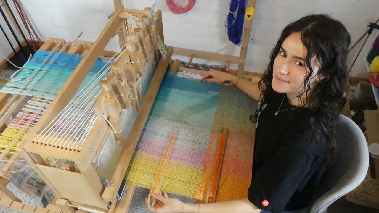 Une jeune femme souriante, passionnée par son tissier métier, travaille sur un métier à tisser en bois. Les fils colorés créent une œuvre vibrante.