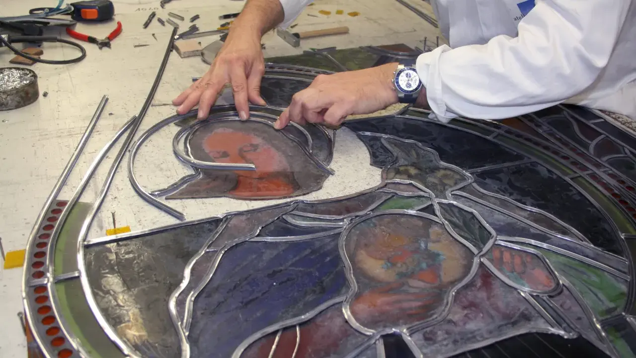 Un artisan assemble un vitrail, joignant des pièces de verre coloré avec du plomb. Le visage d'une Vierge et d'un enfant se dessine.