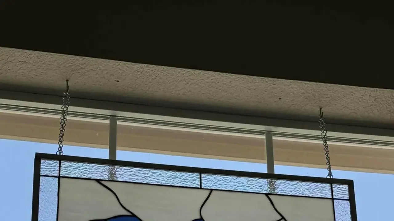 Comment fixer un vitrail sur une fenêtre : un panneau de verre bleu et blanc est suspendu par des chaînes à des crochets fixés au plafond.