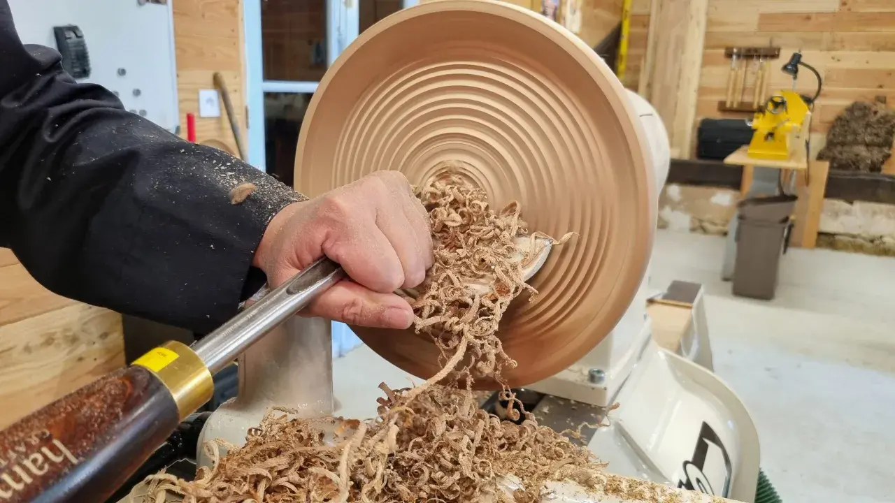 Main d'un artisan façonnant un objet en bois sur un tour. Les copeaux volent, témoignant du travail de tournage sur bois.