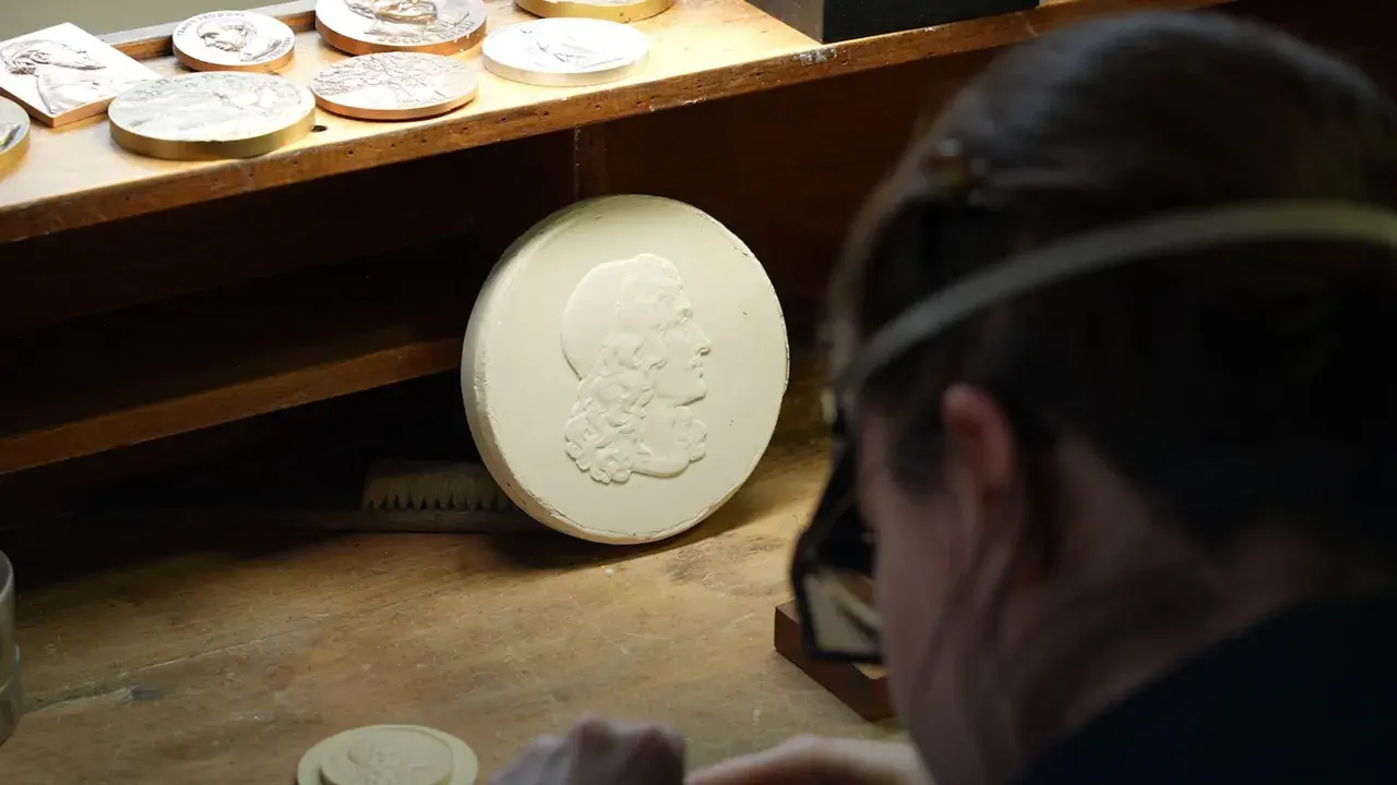 Un graveur métier travaille sur un moule en plâtre orné d'un profil antique. D'autres médailles sont visibles sur l'établi.