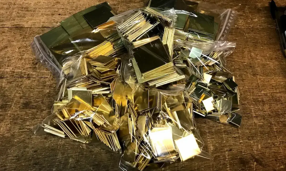 Des petits carrés de laiton brillants, prêts pour un projet de comment décaper du laiton. Ils sont dans des sacs en plastique sur une table en bois.