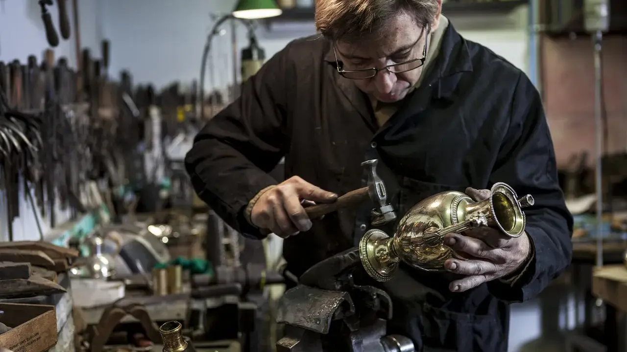 Un orfèvre au travail, martelant délicatement un vase en métal dans son atelier rempli d'outils.