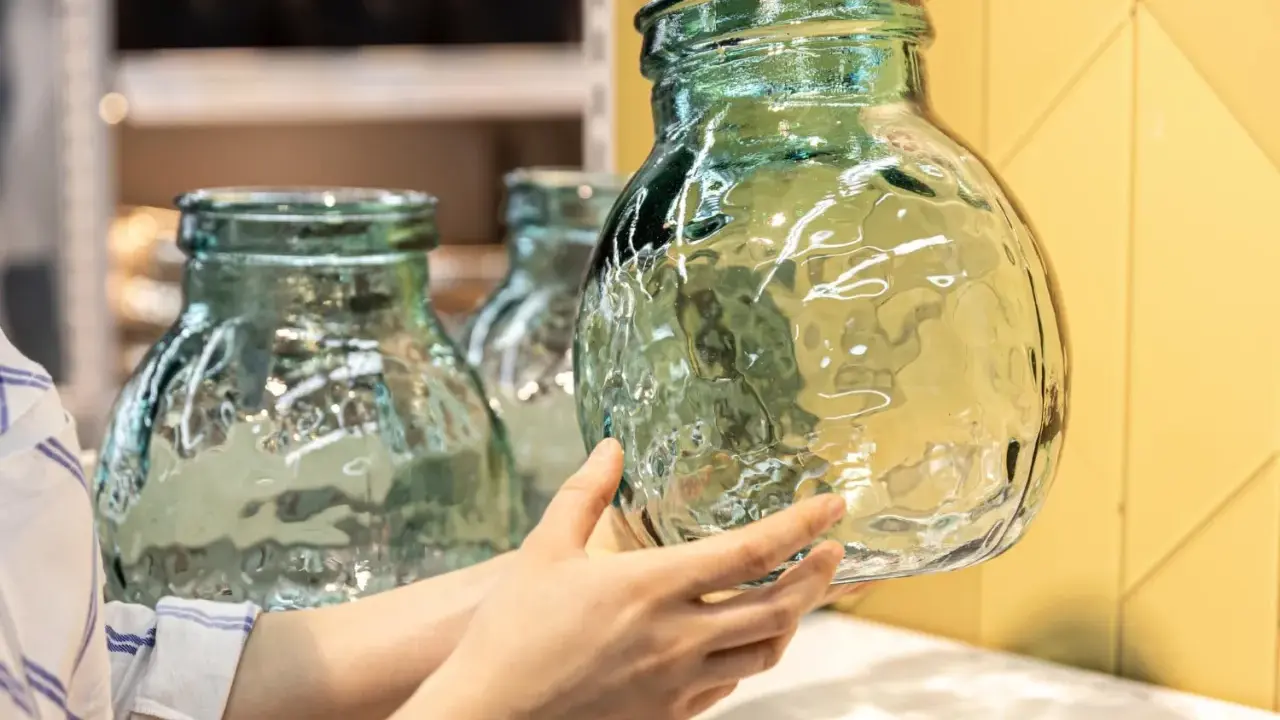 Une main examine un objet en verre texturé, vert pâle, aux reflets ondulés. D'autres objets similaires sont visibles en arrière-plan.
