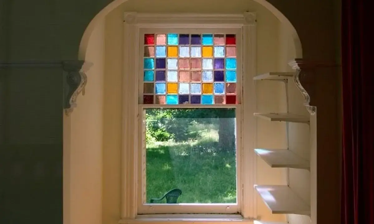 Un vitrail design aux carrés colorés illumine une fenêtre, projetant des reflets joyeux sur l'herbe verdoyante à l'extérieur.