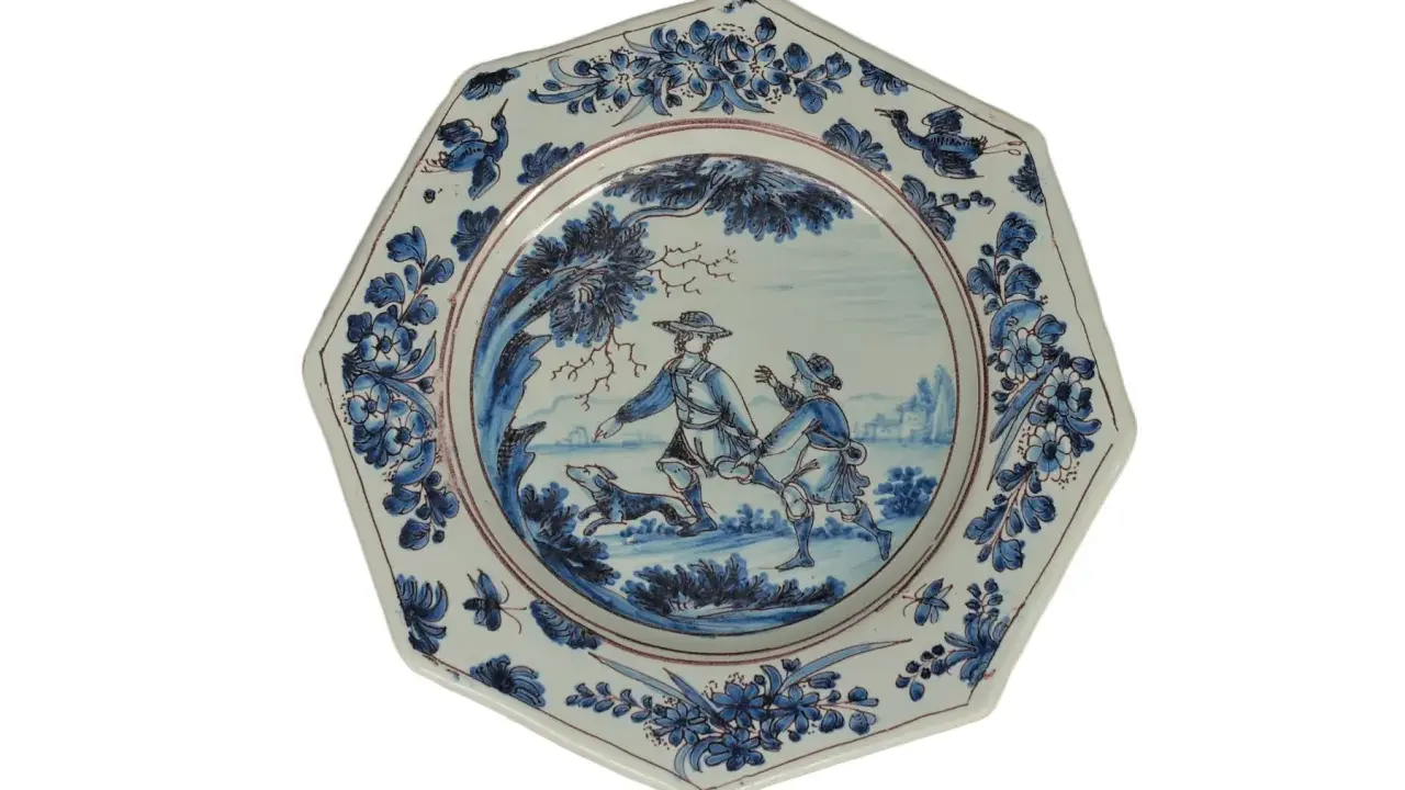 Assiette octogonale en céramique de Marseille, décorée de deux hommes et d'un chien dans un paysage champêtre, bordée de motifs floraux bleus.