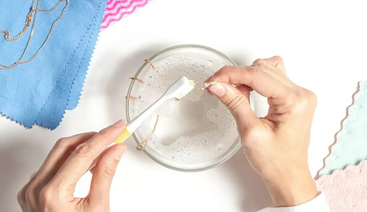 Comment faire briller un diamant : nettoyage d'une bague avec une brosse à dents dans de l'eau savonneuse. Une chaîne en or est posée sur un tissu bleu.
