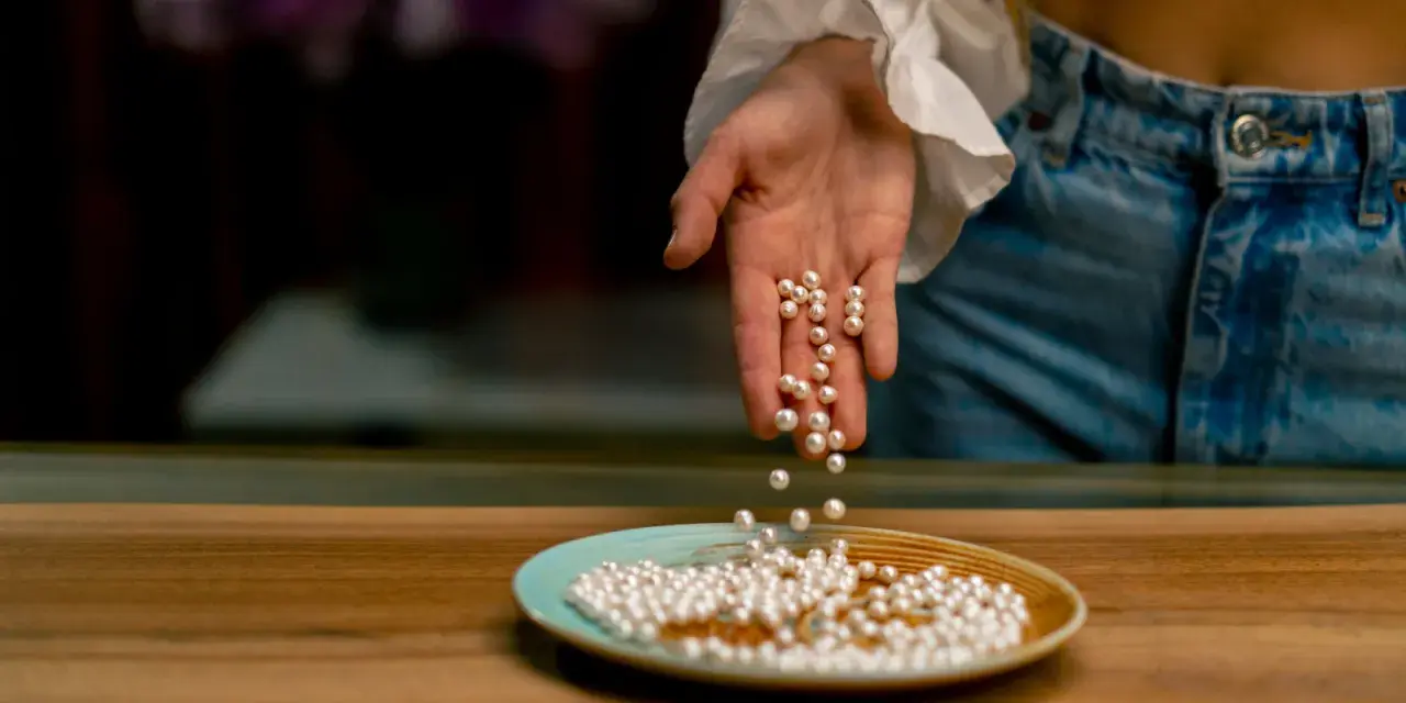 Main d'une personne versant des perles de culture sur une assiette. Apprenez comment nettoyer les perles de culture pour qu'elles brillent.