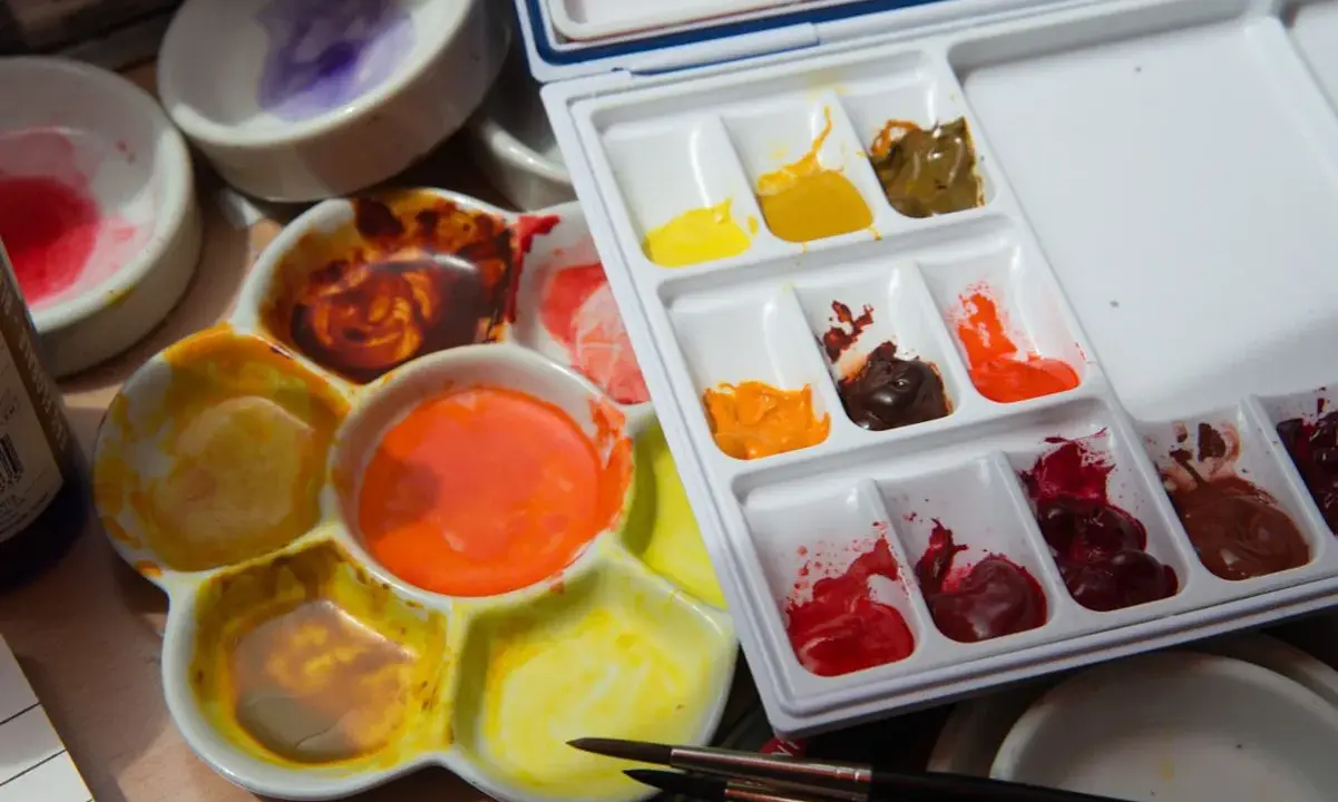 Palette d'aquarelles avec des couleurs vives, dont un mélange de **couleur jaune brun** dans un creux. Des pinceaux sont prêts à l'emploi.