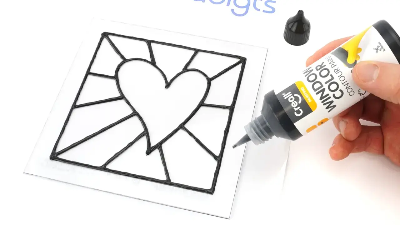 Main noire pour un modèle vitrail en forme de cœur. Un tube de peinture contour est tenu par une main.