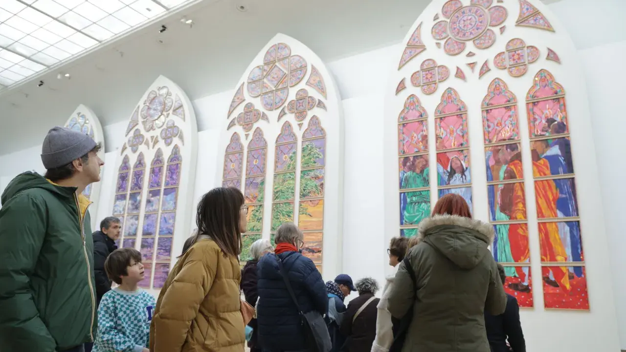 Des visiteurs contemplent de grands vitraux contemporains aux couleurs vives, représentant des scènes et des paysages.