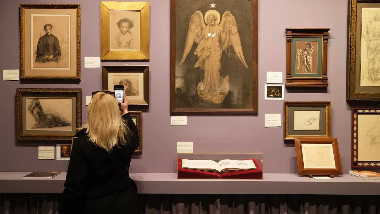 Visite d'un salon du dessin : une femme photographie une œuvre d'art, entourée de nombreux croquis et peintures exposés sur un mur lilas.