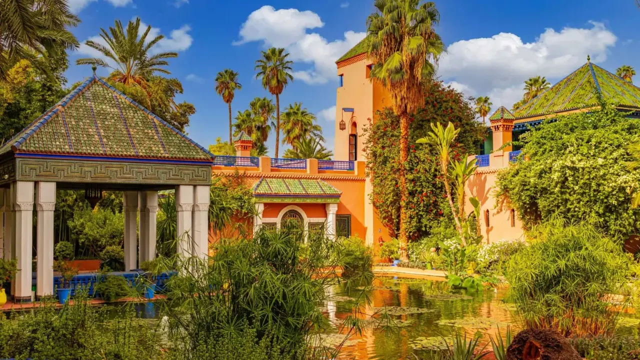 Jardin luxuriant avec un étang reflétant une maison bleue à Marrakech, palmiers et ciel bleu.