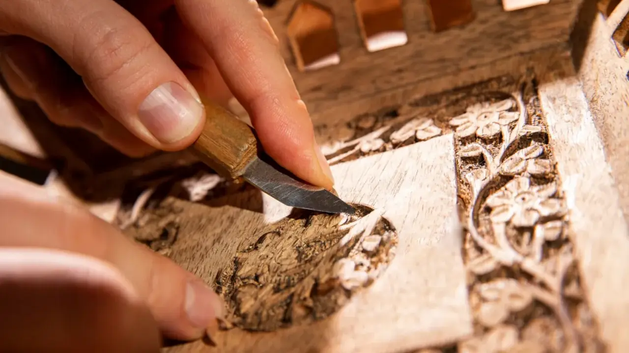Main d'artisan sculptant un détail floral sur un meuble en bois, étape clé pour savoir comment nettoyer un meuble en marqueterie.