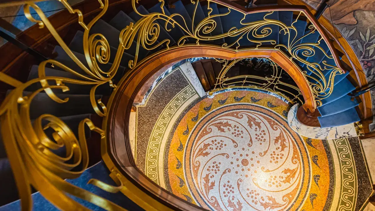 Escalier en colimaçon orné d'une rampe dorée aux volutes végétales, rappelant une fleur art nouveau. Le sol en mosaïque circulaire est un chef-d'œuvre.
