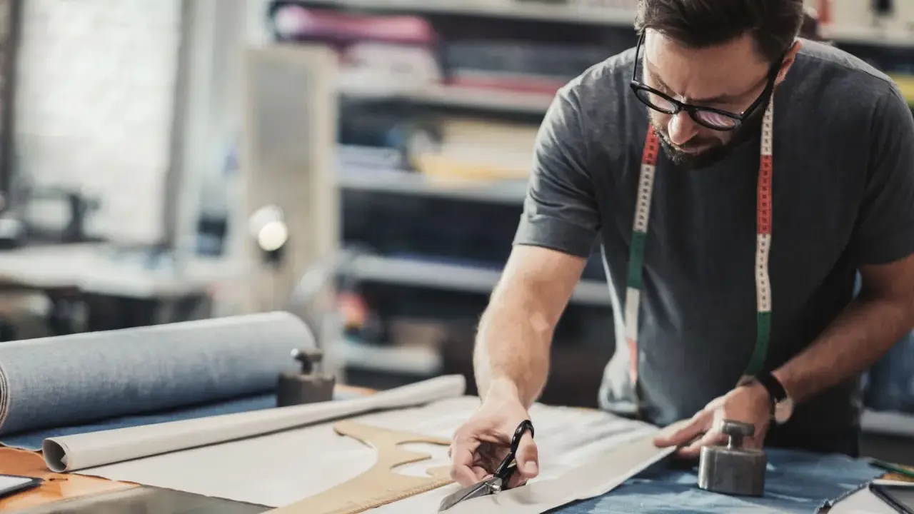 Un artiste textile concentré, lunettes sur le nez, coupe un tissu avec précision dans son atelier.