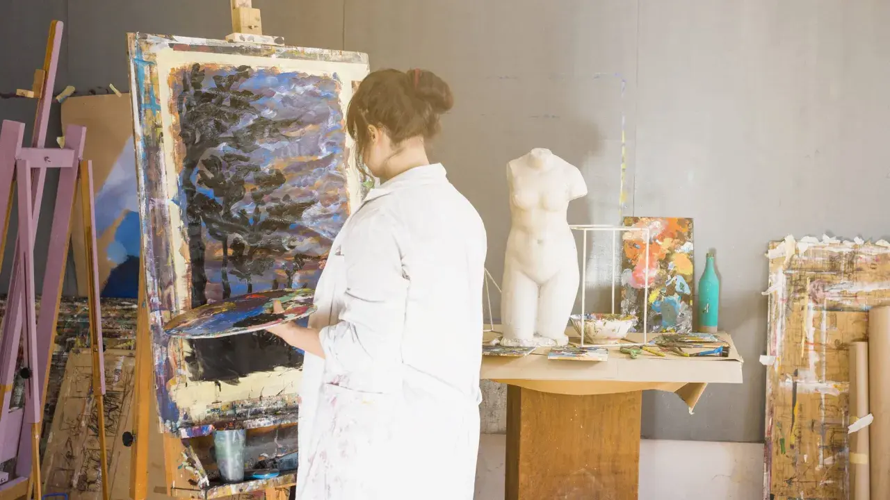 Un artisan artiste, en blouse blanche, tient une palette devant une toile abstraite dans son atelier. Une statue de femme est visible.