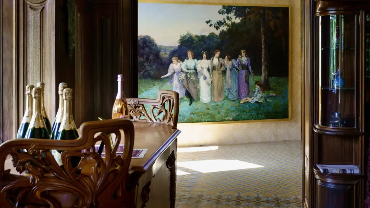 Bouteilles de champagne sur une table en bois sculpté, devant une peinture de femmes dans un jardin, ambiance Villa Mademoiselle Reims.