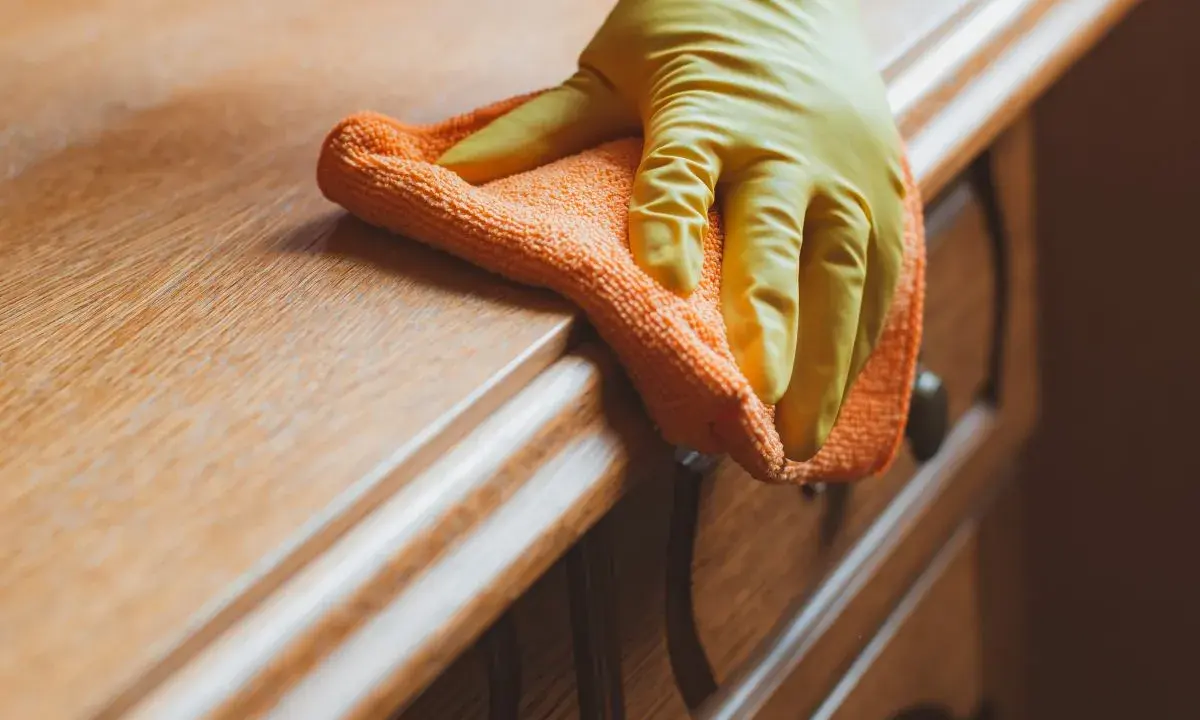 Main d'une personne portant un gant jaune, nettoyant un meuble en bois ciré encrassé avec un chiffon orange.
