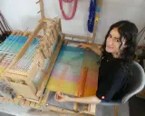 Une jeune femme souriante, passionnée par son tissier métier, travaille sur un métier à tisser en bois. Les fils colorés créent une œuvre vibrante.