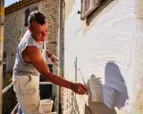 Un homme applique un badigeon blanc sur un mur en pierre avec un large pinceau. Le soleil éclaire la scène, créant des ombres.