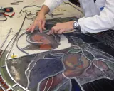Un artisan assemble un vitrail, joignant des pièces de verre coloré avec du plomb. Le visage d'une Vierge et d'un enfant se dessine.