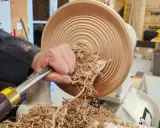 Main d'un artisan façonnant un objet en bois sur un tour. Les copeaux volent, témoignant du travail de tournage sur bois.