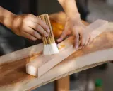 Un artisan ébéniste applique une finition sur un morceau de bois avec un pinceau.