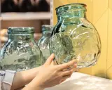 Une main examine un objet en verre texturé, vert pâle, aux reflets ondulés. D'autres objets similaires sont visibles en arrière-plan.