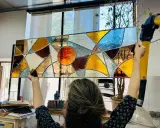 Un artiste admire son œuvre, un vitrail aux couleurs chaudes et géométriques, fruit d'un cours de vitrail.