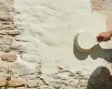 Une main applique une peinture claire sur un mur en pierre enduit de chaux. La question "peut on peindre sur de la chaux" trouve ici sa réponse visuelle.