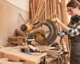 Artisane souriante utilisant une scie à onglet. Elle porte des lunettes et un tablier, des éléments essentiels pour comment protéger une création artisanale.