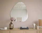 Un miroir vitrail aux formes organiques reflète une pièce épurée. Une fleur séchée dans un vase en verre fumé et une lampe en rotin complètent la scène.