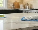 Main d'une personne portant un gant blanc, essuyant un comptoir en pierre naturelle avec un chiffon bleu. Le nettoyage de pierre naturelle à l'intérieur est essentiel pour préserver sa beauté.