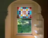 Un vitrail design aux carrés colorés illumine une fenêtre, projetant des reflets joyeux sur l'herbe verdoyante à l'extérieur.