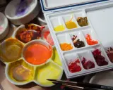 Palette d'aquarelles avec des couleurs vives, dont un mélange de **couleur jaune brun** dans un creux. Des pinceaux sont prêts à l'emploi.