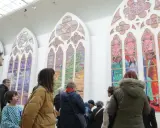 Des visiteurs contemplent de grands vitraux contemporains aux couleurs vives, représentant des scènes et des paysages.
