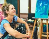 Une artiste rêveuse, les mains tachées de peinture, s'interroge sur comment devenir artisan d'art. Son atelier est rempli de toiles colorées.