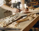 Un artisan qui travaille le bois, concentré sur la précision de ses gestes. Des copeaux de bois jonchent l'établi.