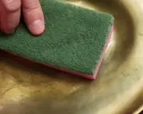 Main tenant une éponge verte et rose, une main frotte un plateau en cuivre jaune pour le nettoyer.