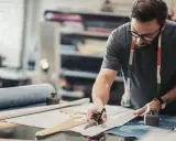Un artiste textile concentré, lunettes sur le nez, coupe un tissu avec précision dans son atelier.