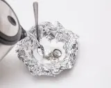 Nettoyage d'argenterie noircie : de l'eau chaude est versée sur des bijoux en argent et une cuillère dans une coupelle en papier aluminium.