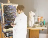 Un artisan artiste, en blouse blanche, tient une palette devant une toile abstraite dans son atelier. Une statue de femme est visible.
