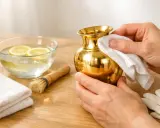 Comment nettoyer du laiton doré : une main frotte un vase doré avec un chiffon blanc. À côté, un bol d'eau citronnée, une brosse et des gants.