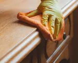 Main d'une personne portant un gant jaune, nettoyant un meuble en bois ciré encrassé avec un chiffon orange.