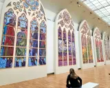 Des vitraux contemporains de Notre-Dame, des œuvres vibrantes composées de multiples panneaux peints, évoquant des scènes humaines et des paysages colorés.