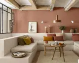 Salon cosy aux murs couleur brun terre de France, banquette en L, coussins ocre et rouille, table basse en bois brut.