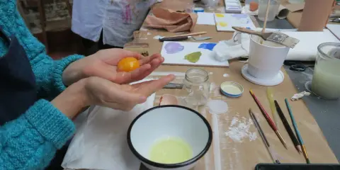 Préparation d'une tempera peinture à l'oeuf : un jaune d'œuf est tenu dans une main au-dessus d'un bol contenant le blanc.