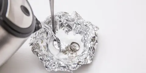 Nettoyage d'argenterie noircie : de l'eau chaude est versée sur des bijoux en argent et une cuillère dans une coupelle en papier aluminium.