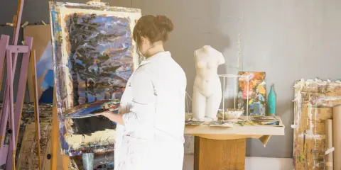 Un artisan artiste, en blouse blanche, tient une palette devant une toile abstraite dans son atelier. Une statue de femme est visible.